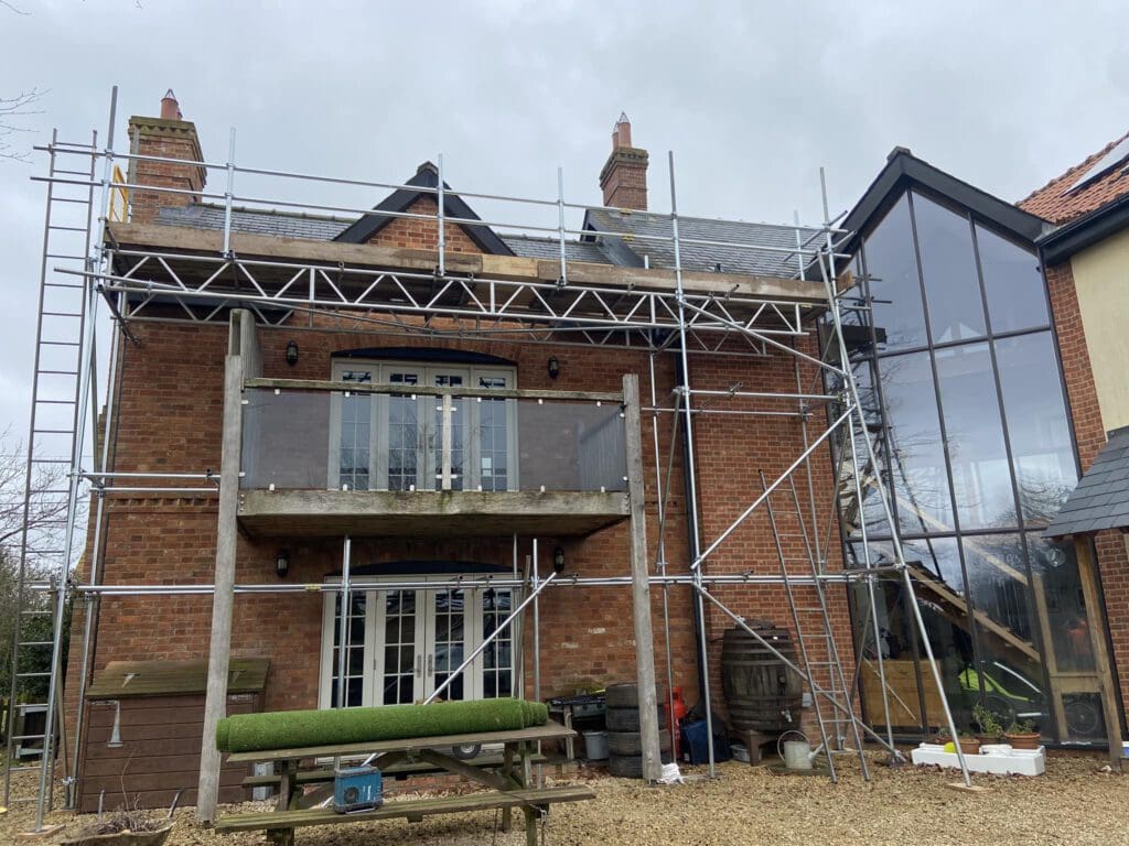 A two-story brick house under construction with scaffolding. There's a large glass extension on the right, and patio doors on both floors. A picnic table with artificial grass on top sits in the foreground, under a cloudy sky.