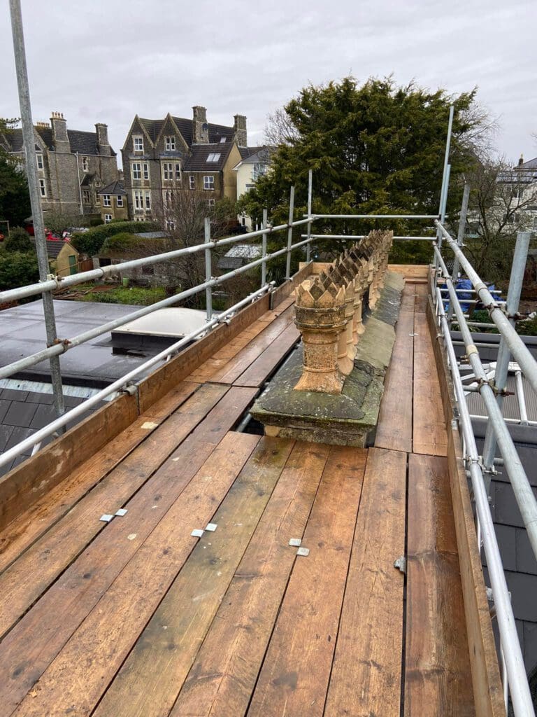 Scaffolding surrounds a rooftop with a wooden platform. Decorative stonework, including ornate chimney pots, is visible along the roof's edge. Houses and trees are in the background under a cloudy sky.