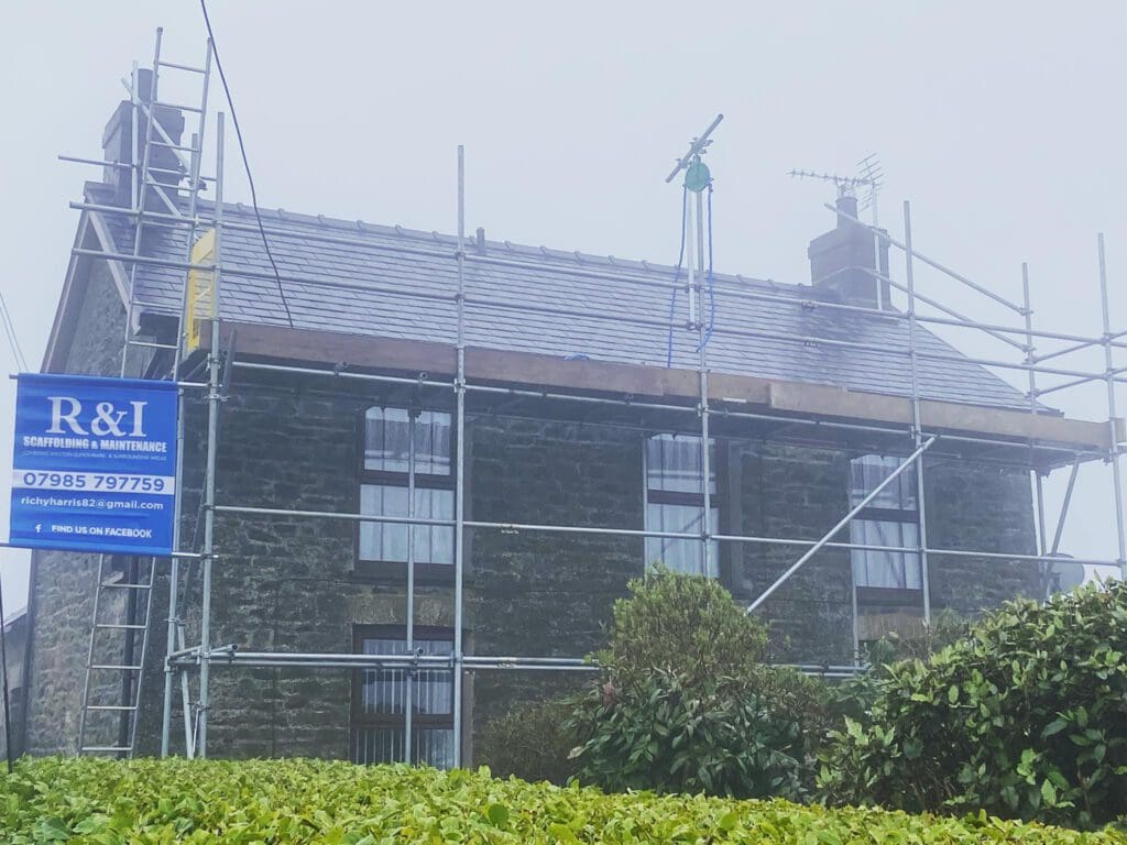 A stone house under renovation is surrounded by scaffolding. The mist adds a hazy atmosphere. A blue sign on the scaffolding displays contact information and a logo. Bushes are visible in the foreground.