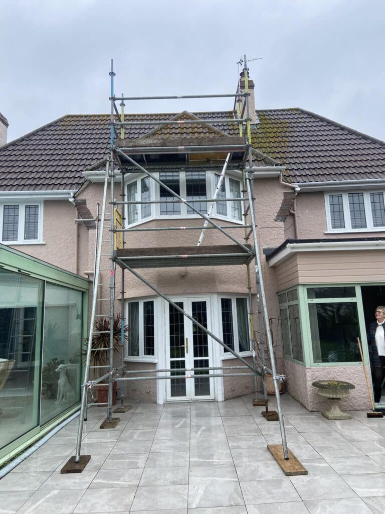 A house with beige exterior and a tiled roof has a scaffolding structure set up in front. The house features large windows and a glass extension on the left. A person stands near a glass door on the right. The ground is paved with light-colored tiles.