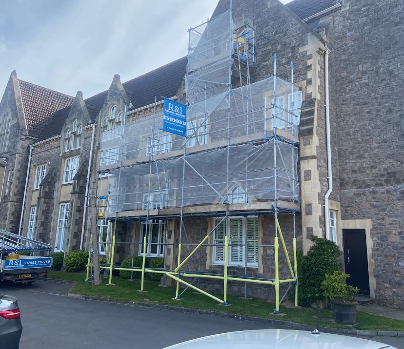 A historic stone building with a sloped roof is partially covered in scaffolding for construction or renovation work. A blue sign with white text is attached to the scaffolding. The surrounding area features a paved driveway and green shrubbery.