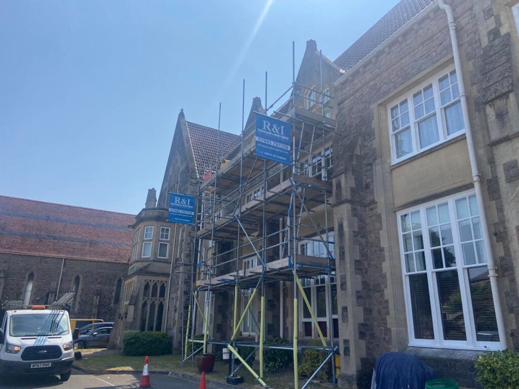 Scaffolding is set up against a large stone building with pointed roofs. A white truck is parked nearby, and traffic cones are visible. The sky is clear, indicating a sunny day. Signs on the scaffolding display "R&T.