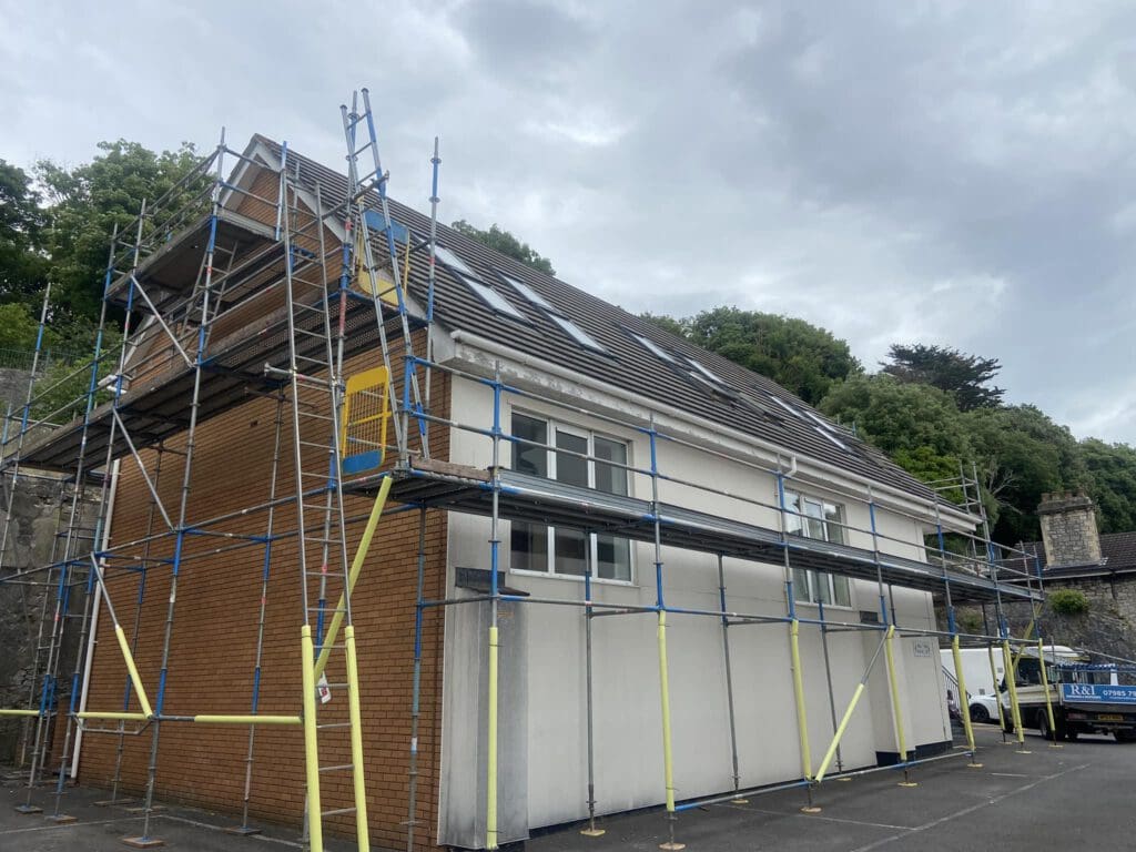 A two-story building is under construction, surrounded by scaffolding. The structure has a combination of brick and white siding. Trees are visible in the background under a cloudy sky.