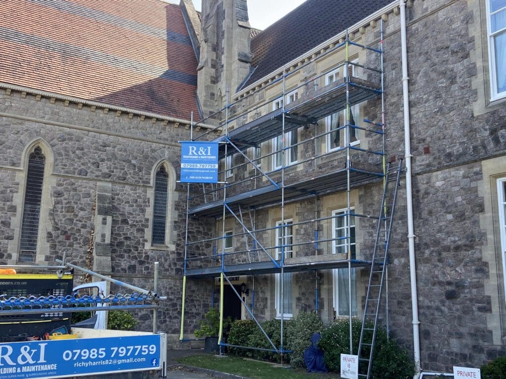 Scaffolding set up against a stone building, with a blue and white "R&I" sign. A van with a company banner is parked nearby. The building features arched windows and is surrounded by some greenery.