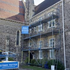 Scaffolding set up against a stone building, with a blue and white "R&I" sign. A van with a company banner is parked nearby. The building features arched windows and is surrounded by some greenery.