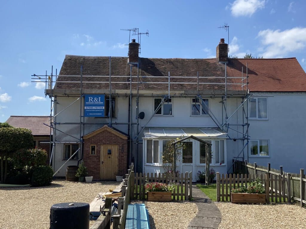 House under renovation with scaffolding on the front facade. Roof tiles are partially removed. A small garden with flowers and a picket fence is visible in the foreground. The sky is clear and sunny.