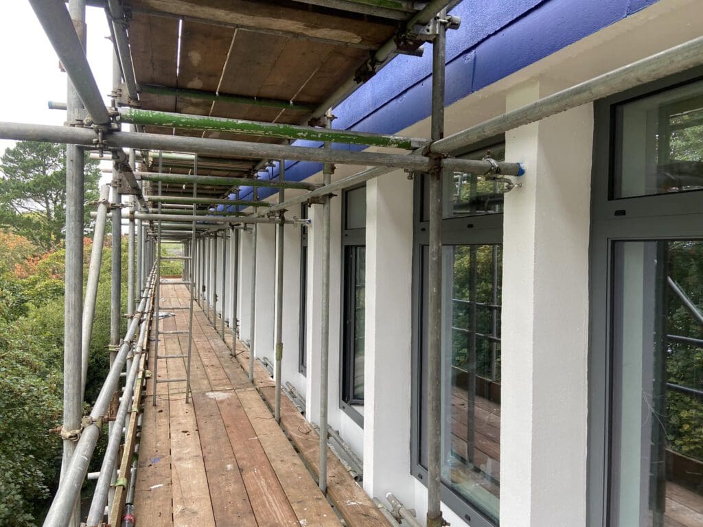 Scaffolding set up along the side of a building with white walls and large windows. The structure features wooden planks and metallic bars, with a blue ledge running above the windows. Trees are visible in the background.