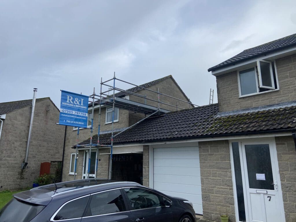 A stone house with scaffolding on the roof, indicating construction work. A blue sign with contact information is attached to the scaffolding. A gray car is parked in the driveway next to a white garage door. The sky is overcast.