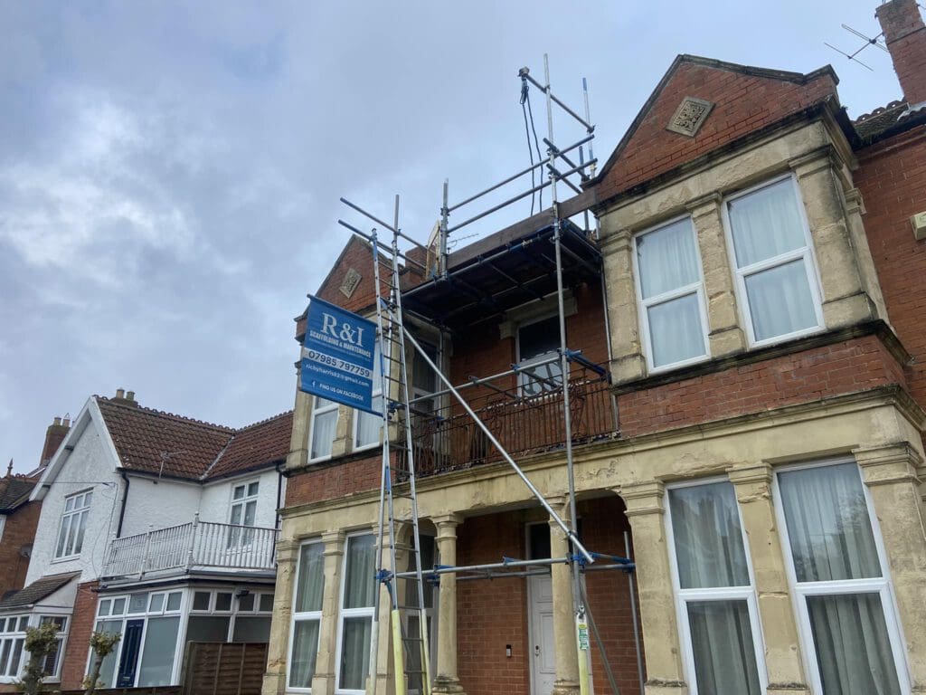 A two-story brick house with scaffolding set up on the front. There's a blue sign on the scaffolding that reads "R&I Contractors." The sky is cloudy, and there are neighboring houses visible on the left.