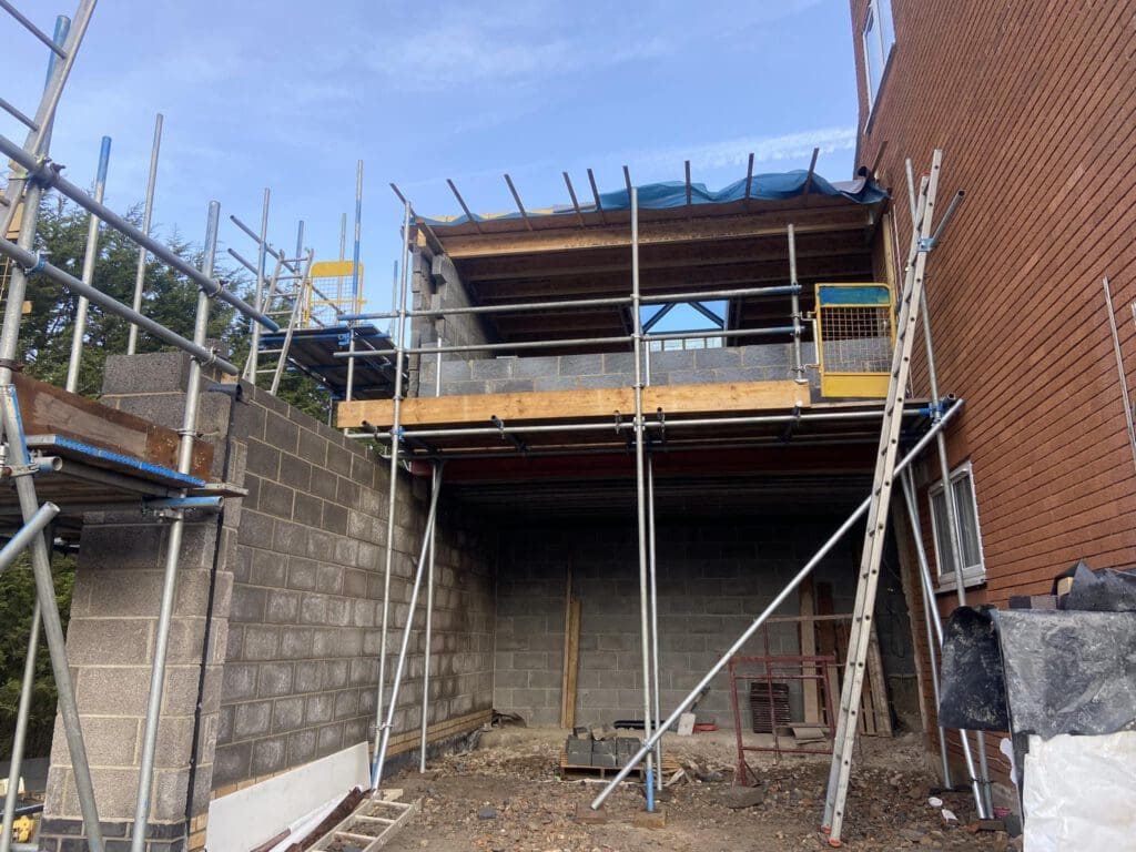 A partially constructed building with scaffolding and brick walls. The structure is connected to an existing brown brick building. The ground is covered with construction materials and debris, and the sky is clear.