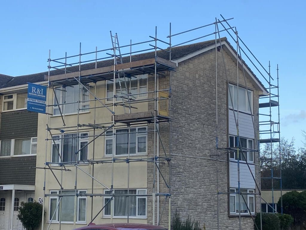 A two-story house with scaffolding on the front and side, indicating construction or renovation work. The building has a mix of brick and siding, and the sky is clear. A sign with "R&I" is attached to the scaffolding.