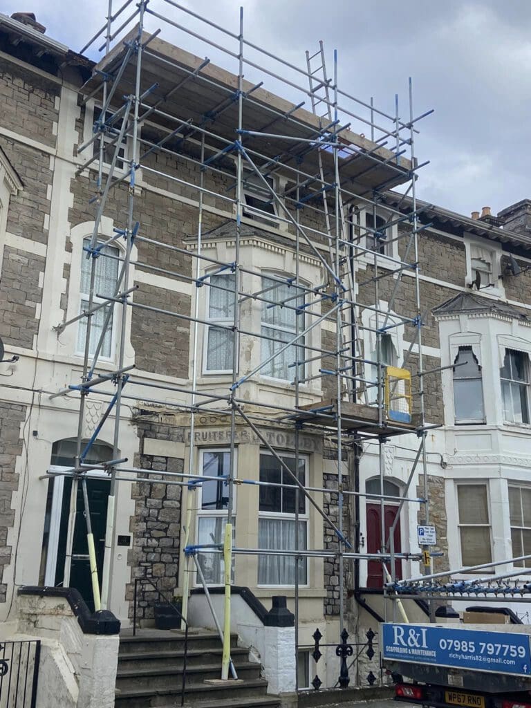 A residential building with scaffolding covers the front, indicating construction or renovation work. A blue truck with company information is parked in front. The house has a stone facade and large windows. The sky is overcast.