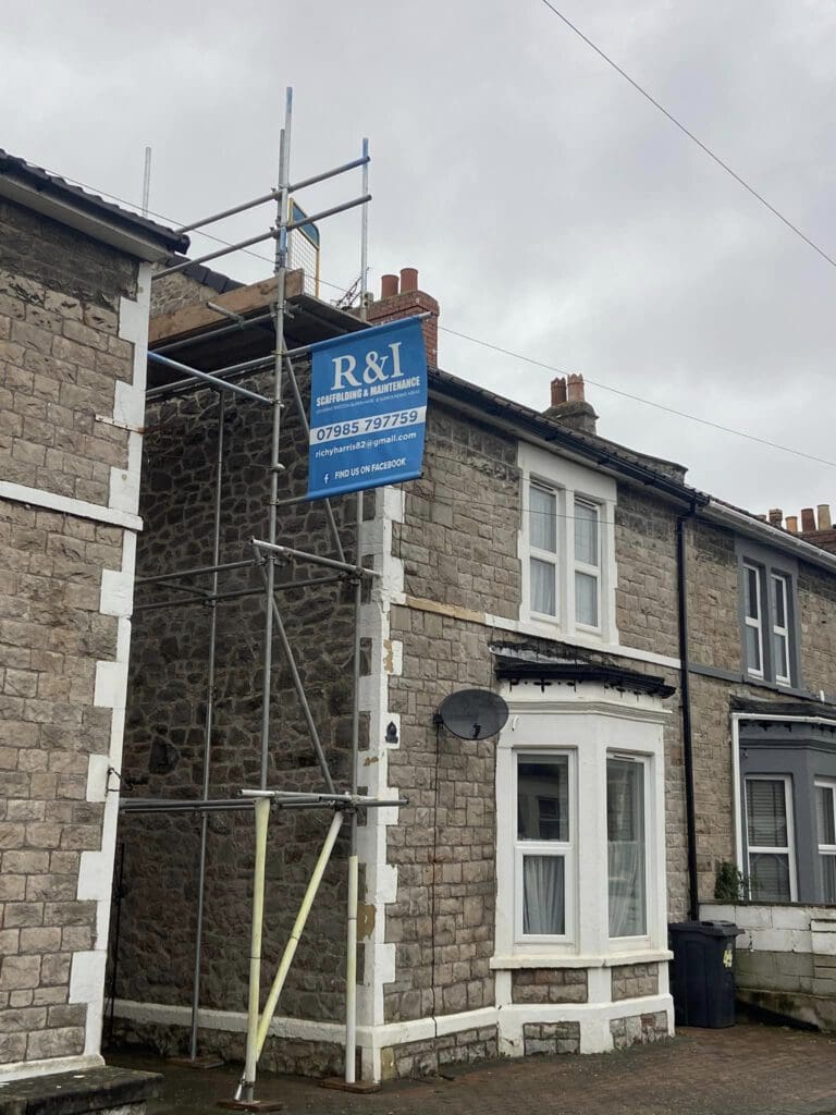 A stone terraced house with scaffolding on one side, featuring a sign for R&I Scaffolding Maintenance. The house has white-trimmed windows and a satellite dish. The sky is overcast.