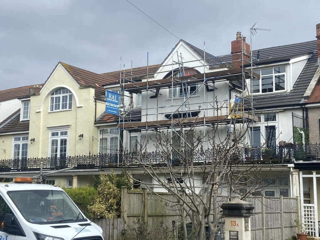 A row of houses with a two-story home under construction. Scaffolding and building materials are visible. A white van is parked in front of the houses. The sky is overcast, and a leafless tree is in the foreground.