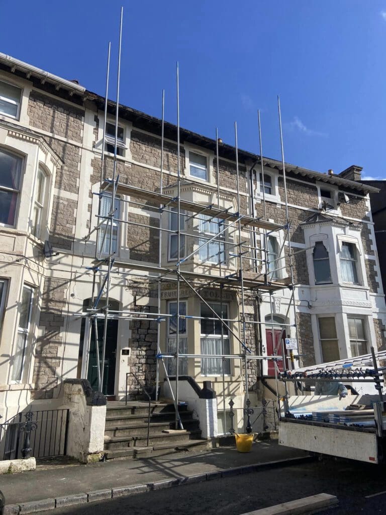 Scaffolding is set up in front of a row of Victorian-style terraced houses. The structure is partially assembled on the facade of one house, with metal poles and platforms visible. The sky is clear and blue.