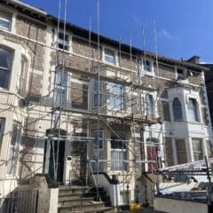 Scaffolding set up in front of a stone-facaded building with a stepped entrance. A yellow bucket is on the ground, and blue skies are overhead.