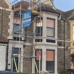 A two-story house under renovation has scaffolding on the front. There's an R&I building services sign displayed on the scaffolding. The building has a stone facade and two large windows covered with orange material. A partial view of a car is in the foreground.