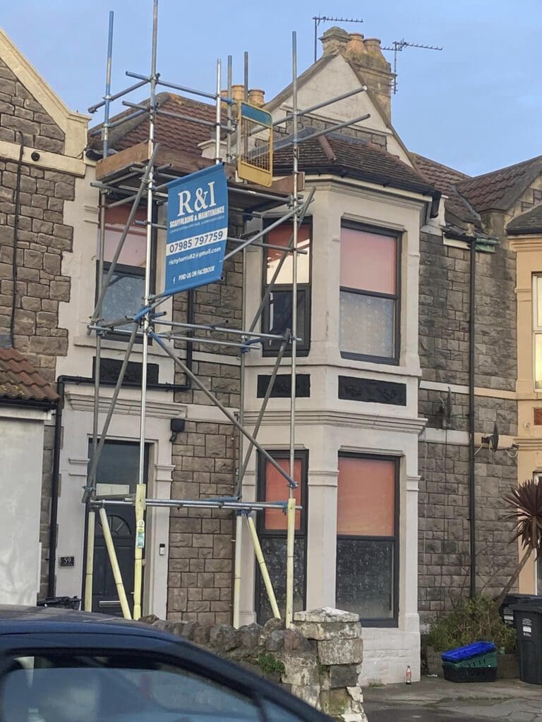 A two-story house under renovation has scaffolding on the front. There's an R&I building services sign displayed on the scaffolding. The building has a stone facade and two large windows covered with orange material. A partial view of a car is in the foreground.