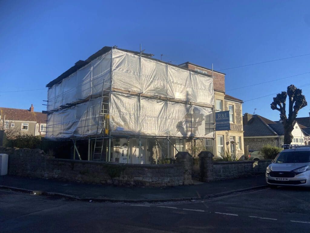 A two-story building under renovation is covered in scaffolding and white tarps. A construction company's sign is visible on the right. A parked car is on the street nearby. The scene is set against a clear blue sky.