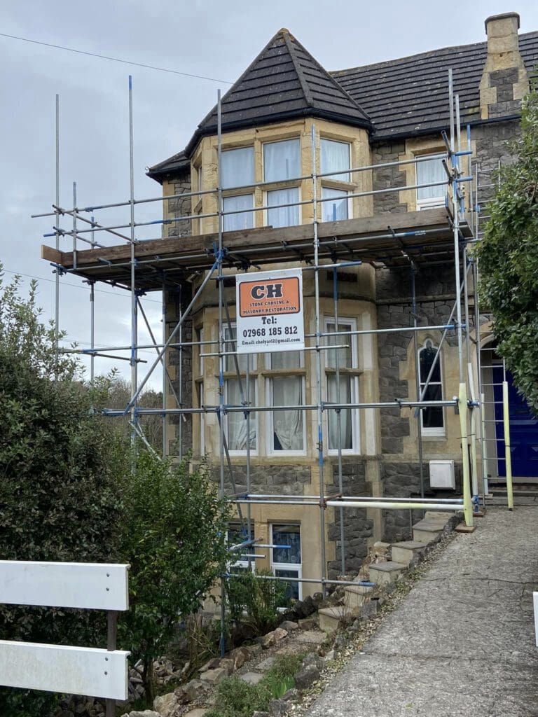 A stone house undergoing renovations, with scaffolding covering the front. A sign on the scaffolding displays the initials "CH" and a phone number. The house has large windows, and the sky is overcast. Nearby, there are green shrubs and trees.