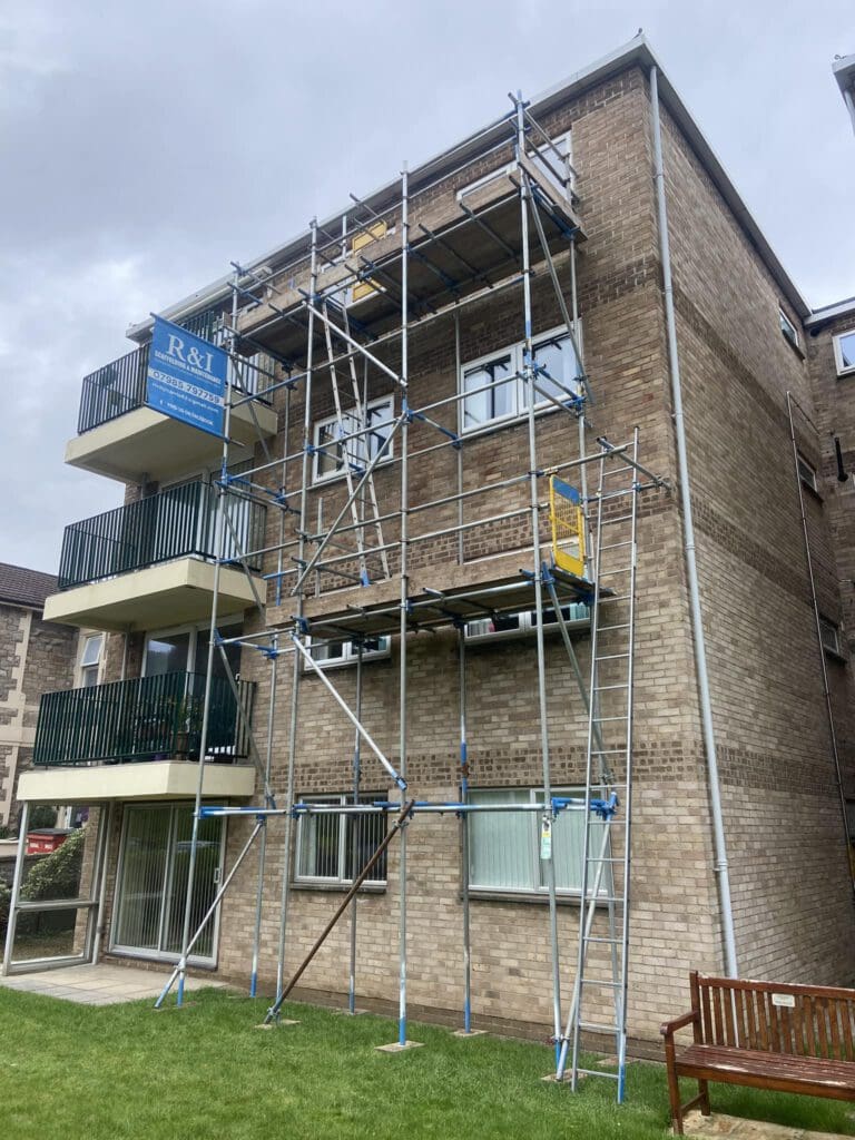 A brick apartment building with scaffolding set up on the side. Balconies with green railings are visible, and a sign on the scaffolding reads "R&I Building Co." A wooden bench is on the grass in the foreground. The sky is overcast.