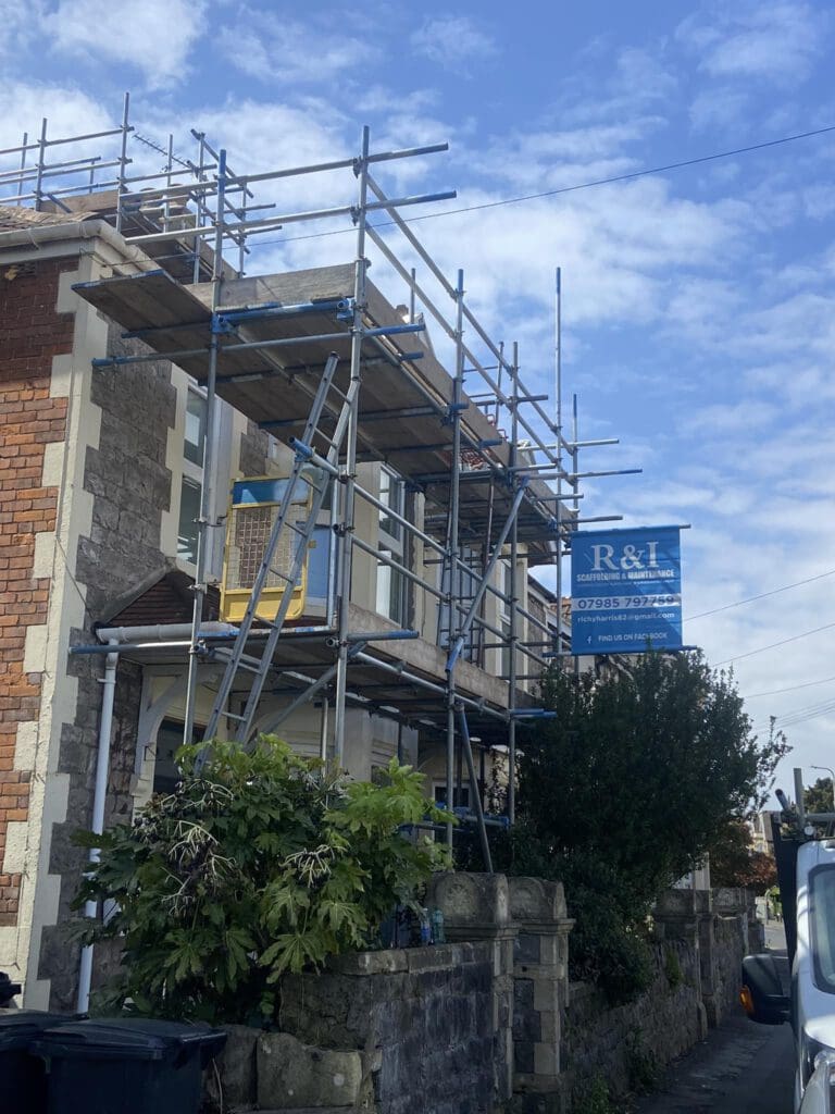 A house under renovation with scaffolding covering the front. A sign with "R&I" is attached to the scaffolding. The sky is partly cloudy. A van is parked on the right, and bushes are along the stone wall in front of the house.