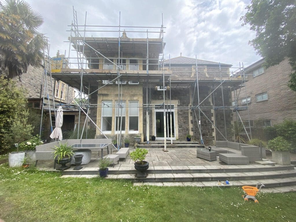 A large house under renovation, surrounded by scaffolding. The building has stone walls and multiple windows. There are outdoor seating and potted plants on the patio, with a grassy lawn and a small toy truck in the foreground.