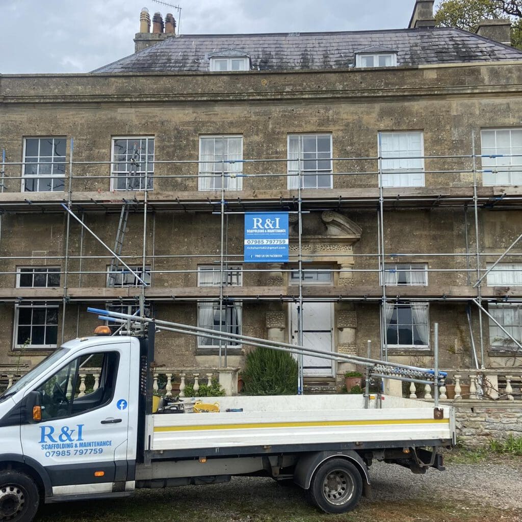 Scaffolding covers the front of an old stone building under renovation. A white truck with "R&I Scaffolding & Maintenance" is parked in front. The building has multiple windows and a central door, with grass and gravel in the foreground.