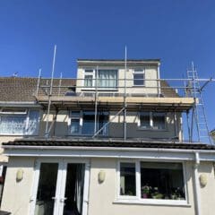 A two-story house with beige walls has scaffolding set up around the upper section. The house has a partially tiled roof and white-framed windows. A clear blue sky is overhead, and there's a garden area visible at the front.