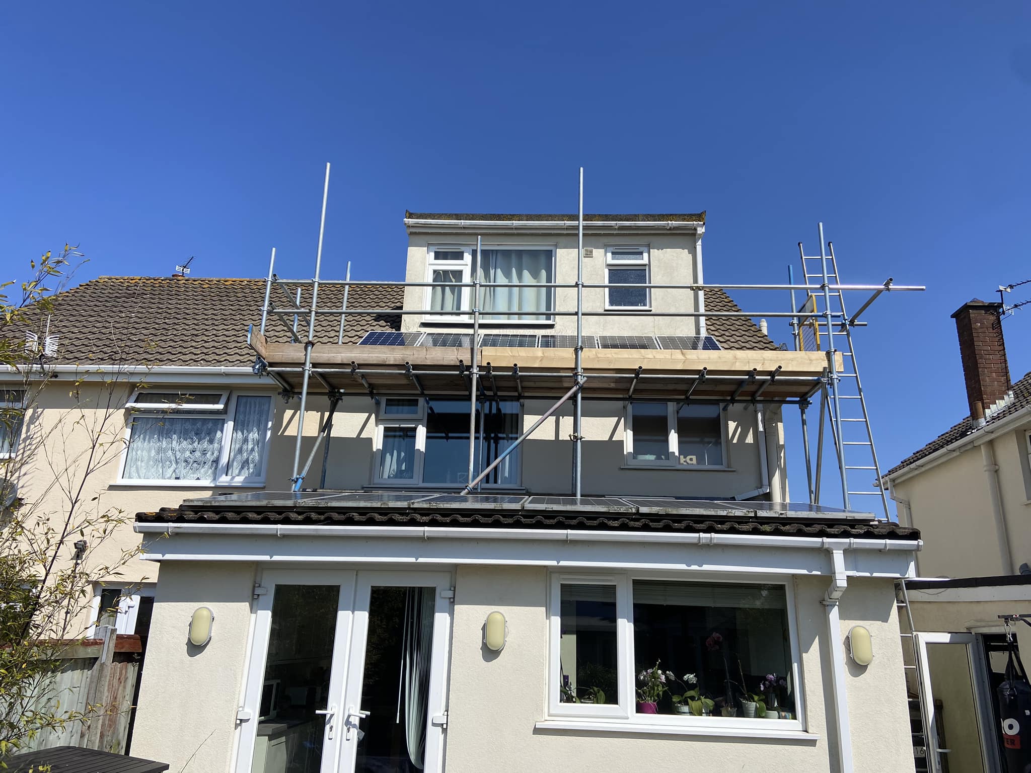 A two-story house with beige walls has scaffolding set up around the upper section. The house has a partially tiled roof and white-framed windows. A clear blue sky is overhead, and there's a garden area visible at the front.