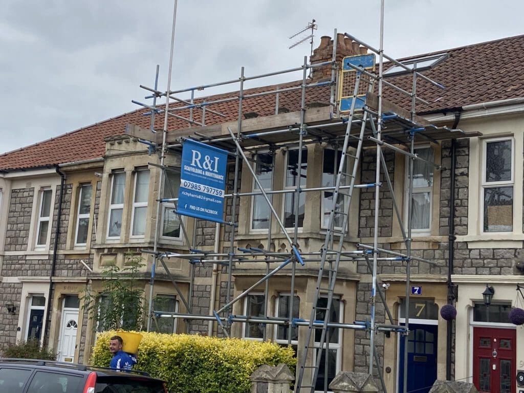 A stone terraced house with scaffolding in front, supporting roof repairs. A sign reads "R&I." A worker stands nearby against a hedge. The sky is overcast.