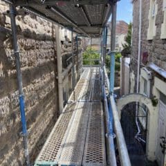 A metal scaffolding walkway set between two stone buildings, offering a view of a garden area at the end. The metal planks have perforations for grip. The sky is clear and the scene is well-lit by daylight.