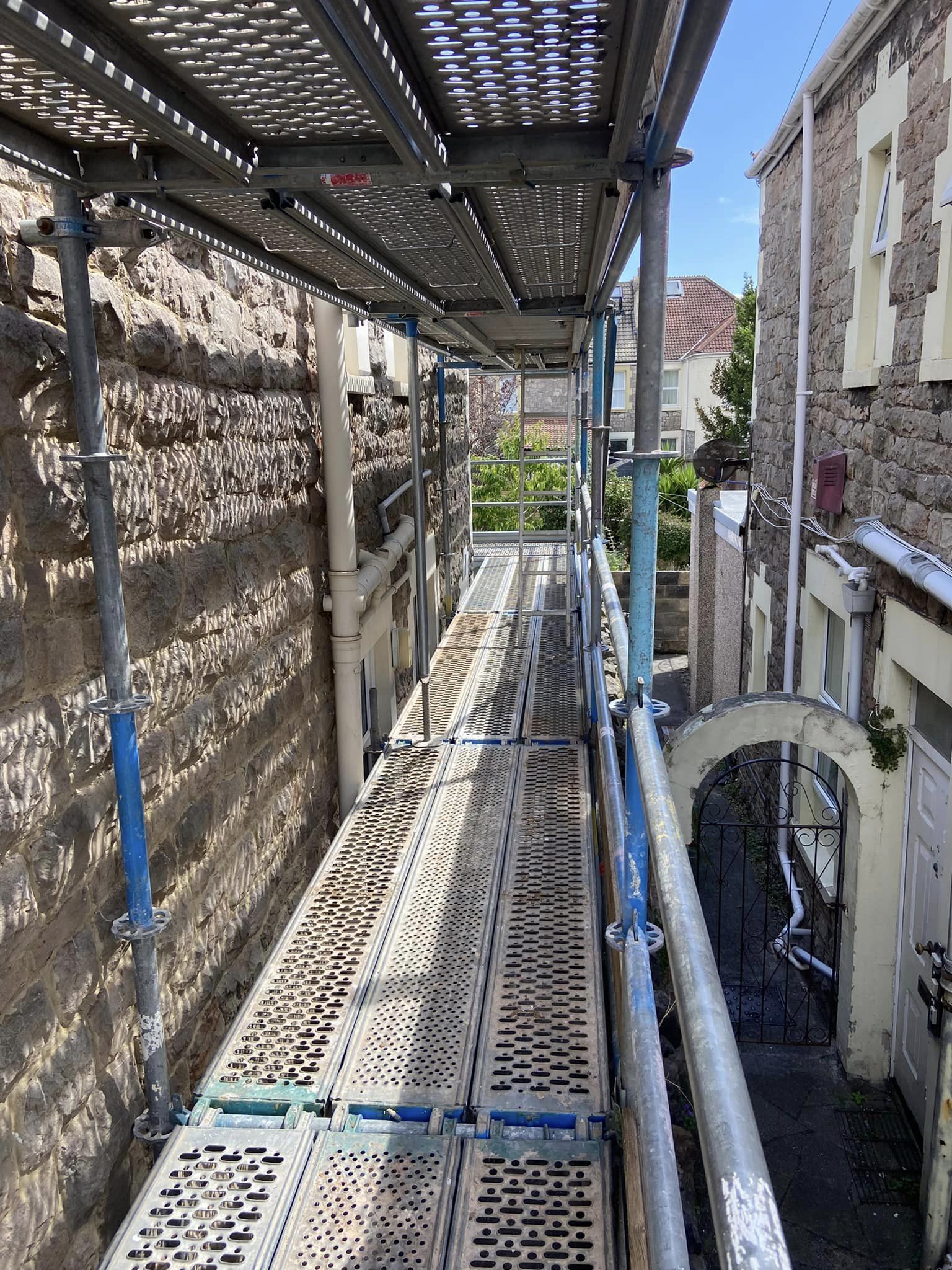 A metal scaffolding walkway set between two stone buildings, offering a view of a garden area at the end. The metal planks have perforations for grip. The sky is clear and the scene is well-lit by daylight.