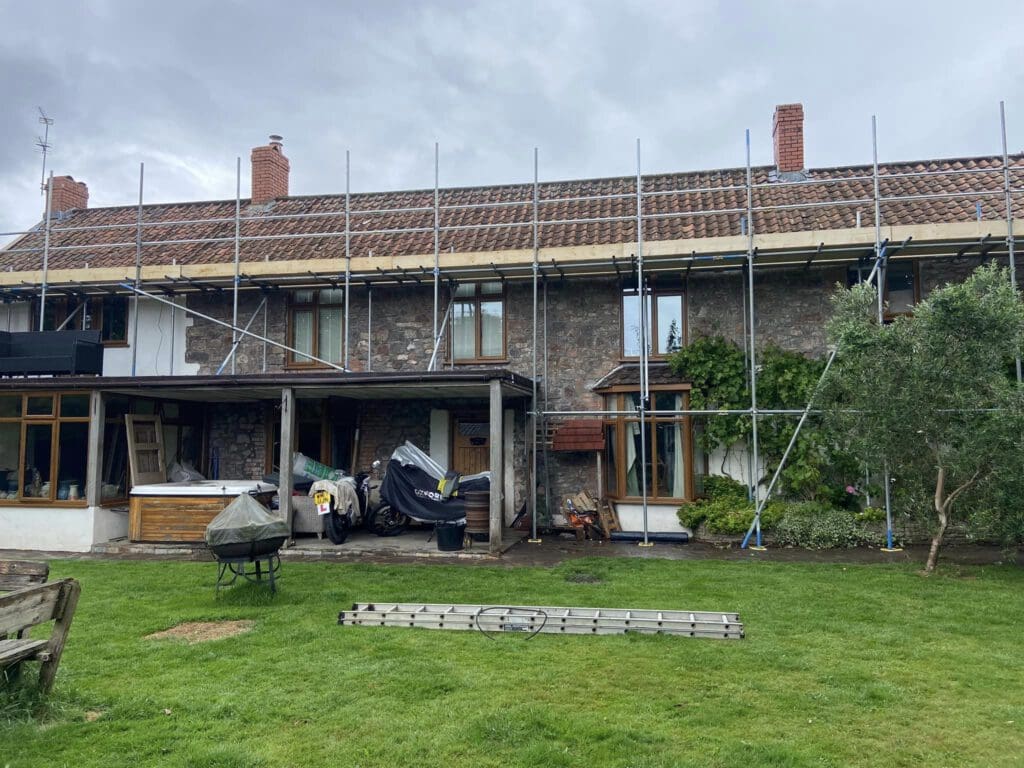 A two-story house with stone walls and a tiled roof is under construction. Scaffolding is set up along the length of the roof. Various building materials and covered items are on the lawn, along with a ladder. The sky is overcast.