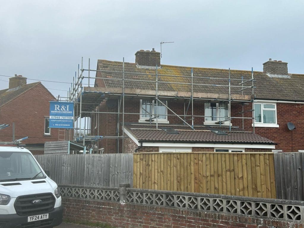 A two-story brick house with scaffolding on the roof, which shows some moss. A sign with "R&I" is attached to the scaffolding. A white van is parked in front of the house next to a wooden fence and a brick wall.