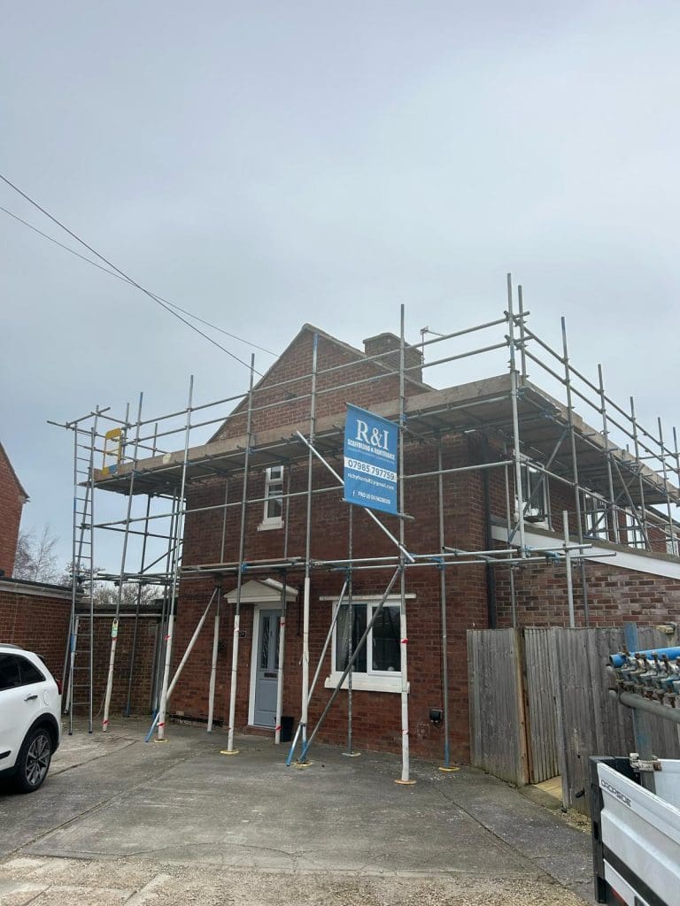 A brick house under renovation, surrounded by metal scaffolding. A sign with "R&I" is visible on the scaffolding. A white SUV is parked in the driveway. The sky is overcast.