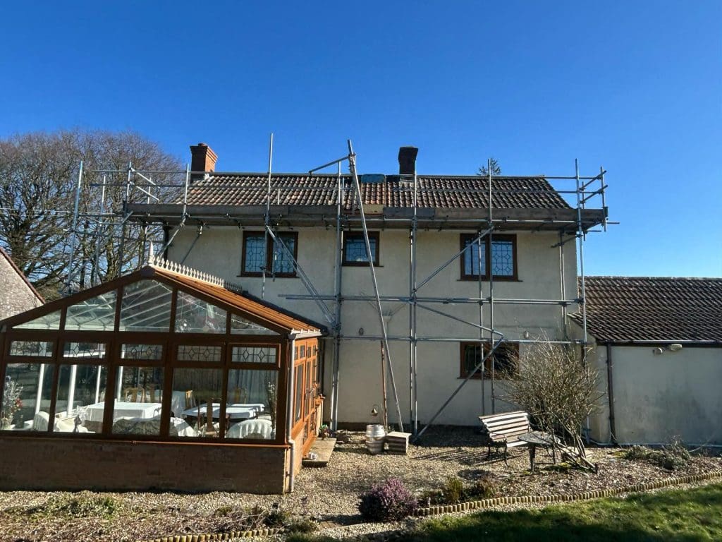 A two-story house under renovation with scaffolding against it. The house has a tiled roof and large windows. In the foreground, there's a glass conservatory, a bench, and some garden plants. The sky is clear and blue.