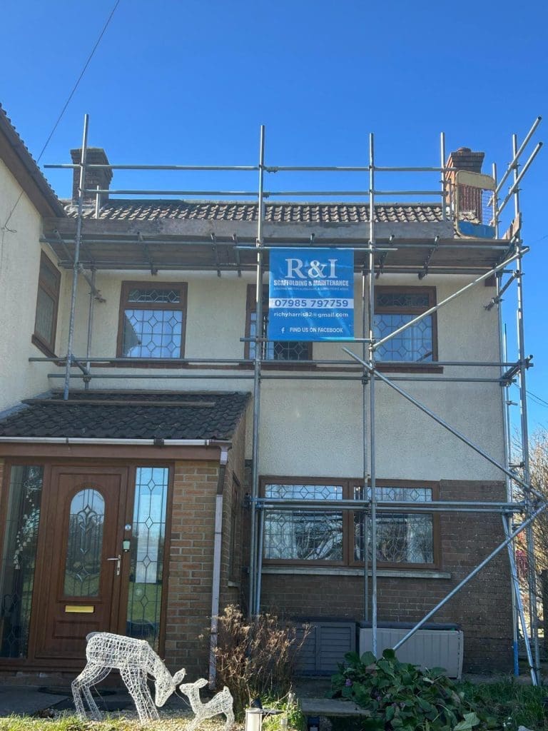 A two-story house with a brown door and several windows is partially covered in scaffolding on the right side. A construction banner is attached to the scaffolding. A white decorative reindeer stands in the yard. The sky is clear and blue.