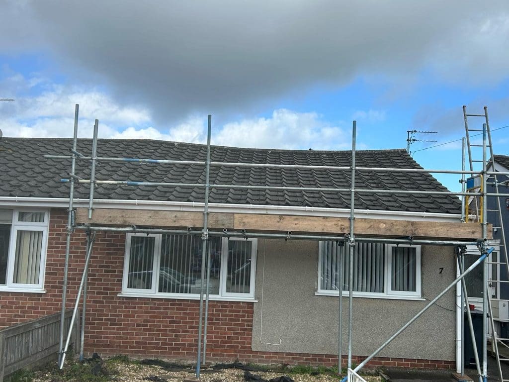 A single-story brick and stucco house with a grey tiled roof is undergoing renovations. Scaffolding is set up along the front, and a ladder is leaning against the roof. The sky is partly cloudy.