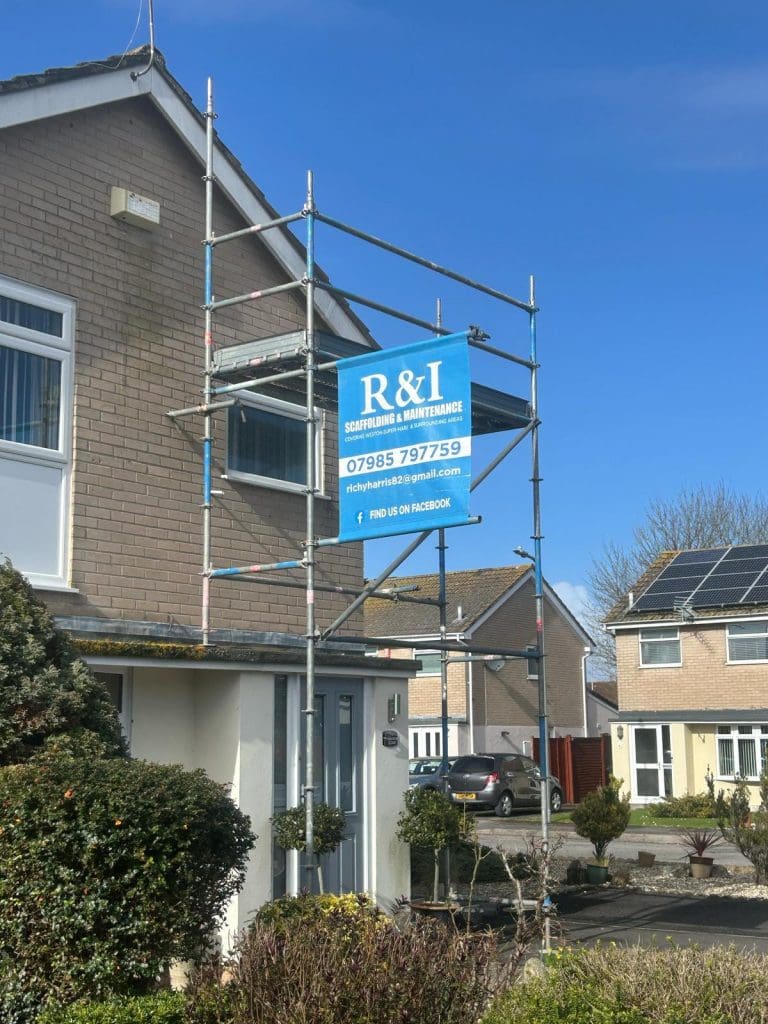 A two-story house with a brick facade has scaffolding on the side. A blue sign on the scaffolding reads "R&I Scaffolding & Maintenance" with contact details. The sky is clear and several houses are in the background.