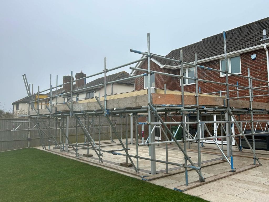 A construction site with metal scaffolding erected next to a two-story brick house. The scaffolding is set on a paved area, and a lawn is in the foreground. The sky appears overcast.