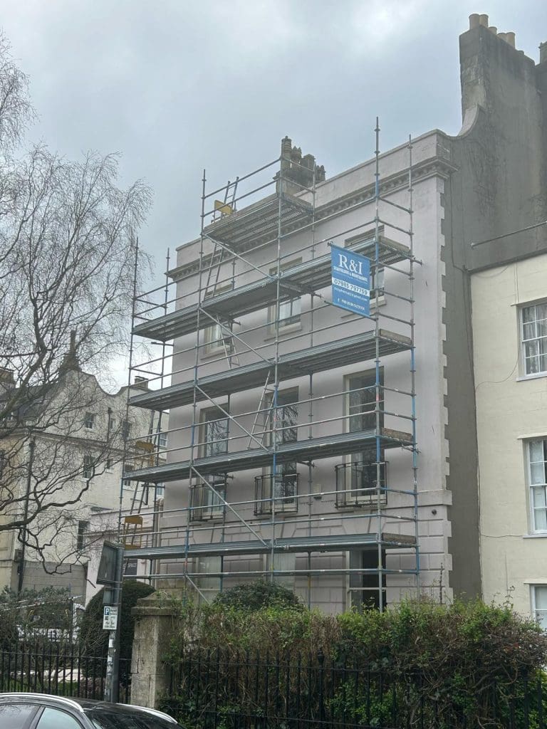 A multi-story building covered in scaffolding, indicating construction or renovation work. The structure is in a residential area with trees and a cloudy sky overhead. A sign on the scaffolding reads "R&I BUILDERS.