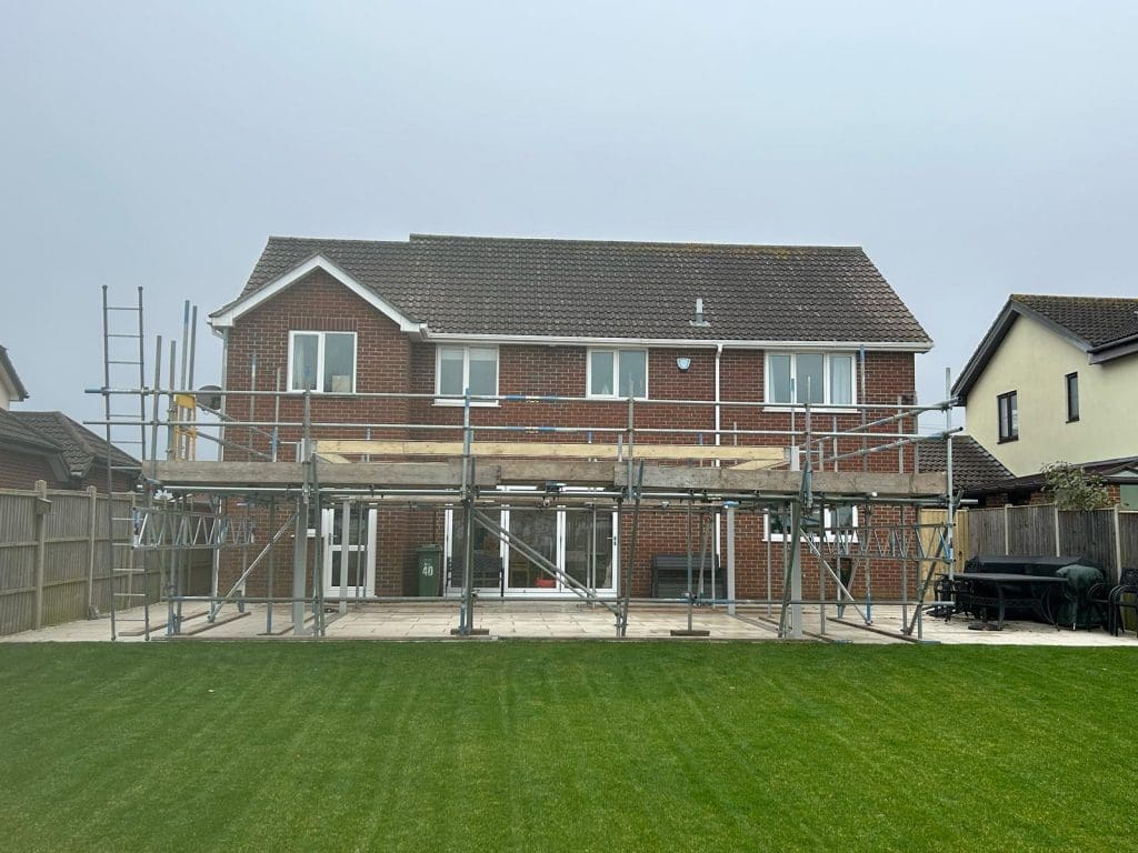 A two-story brick house with scaffolding on the rear exterior, indicating renovation or construction work. The building has a gabled roof and large windows. A green lawn is in the foreground, and neighboring houses are visible.