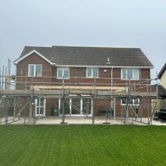 A two-story brick house with scaffolding on the rear exterior, indicating renovation or construction work. The building has a gabled roof and large windows. A green lawn is in the foreground, and neighboring houses are visible.